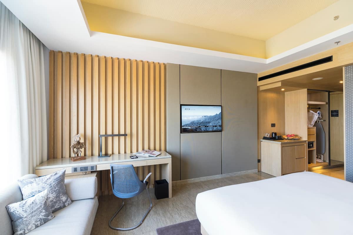 A modern hotel room with a bed, a desk, a chair, a TV, and a kitchenette.