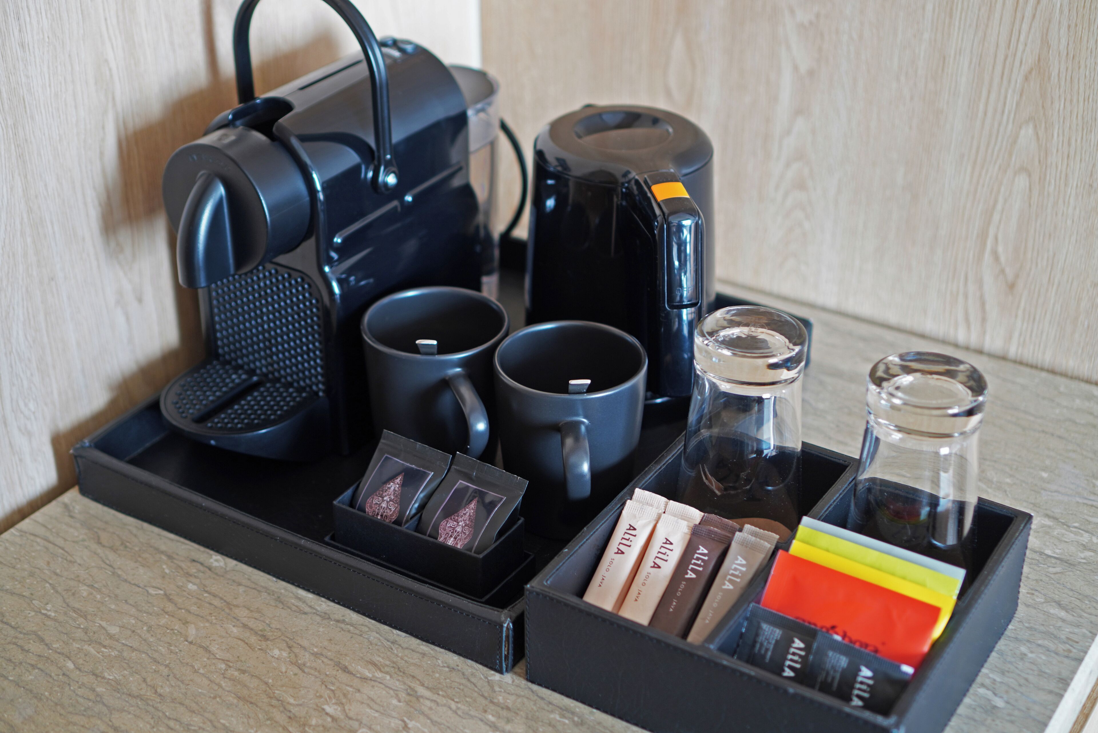Una estación de café con una cafetera, dos tazas, un vaso y una bandeja con sobrecitos de café.