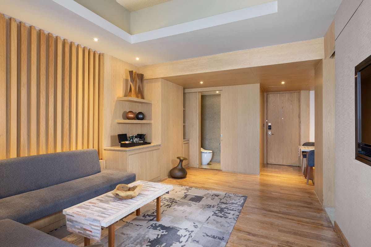 A modern living room with a grey sofa, a coffee table, and a wooden floor. There is a wooden wall with vertical slats and a small shelf with decorative items. A door leads to a bathroom.