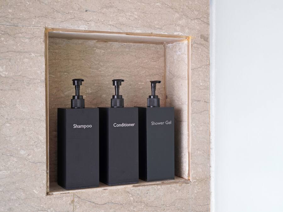 Three black bottles labeled shampoo, conditioner, and shower gel in a marble wall niche.