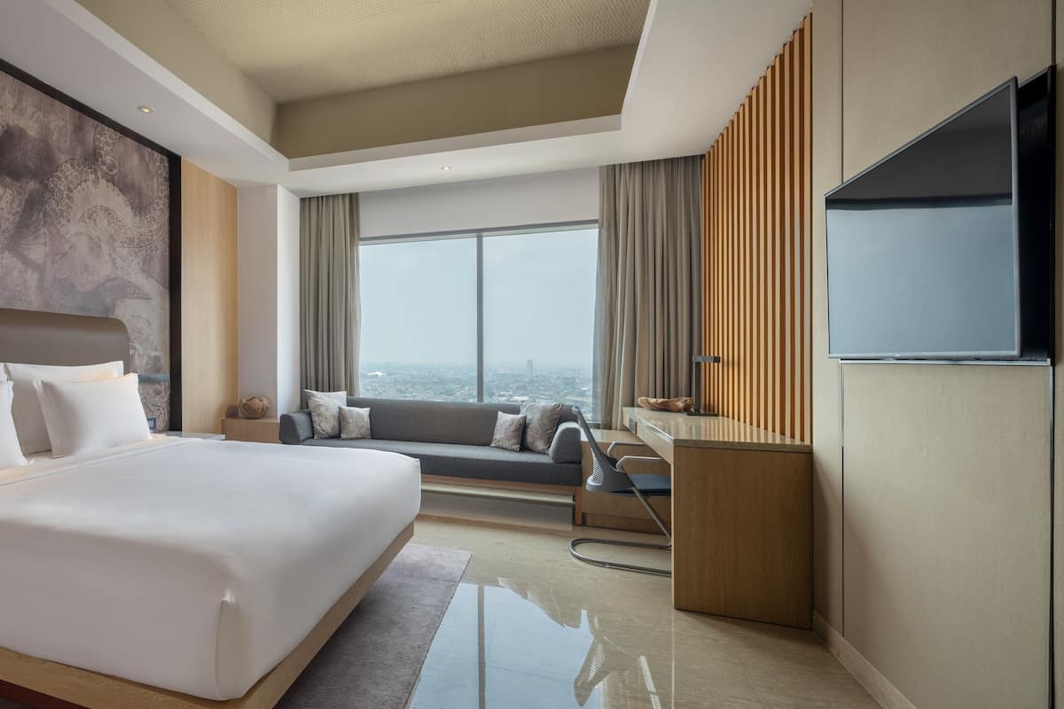 A modern hotel room with a large bed, a sofa, a TV, and a city view.