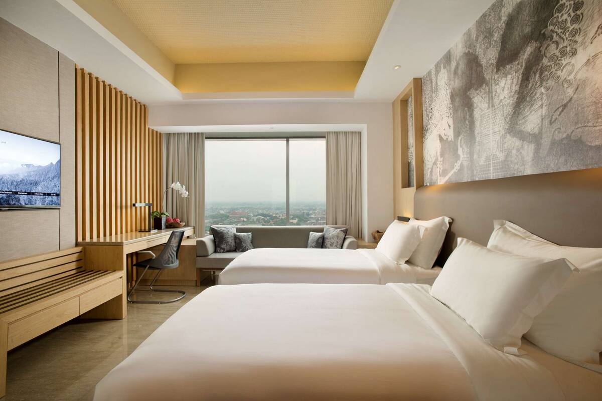 A hotel room with two beds, a desk, a chair, a TV, and a large window with a city view.