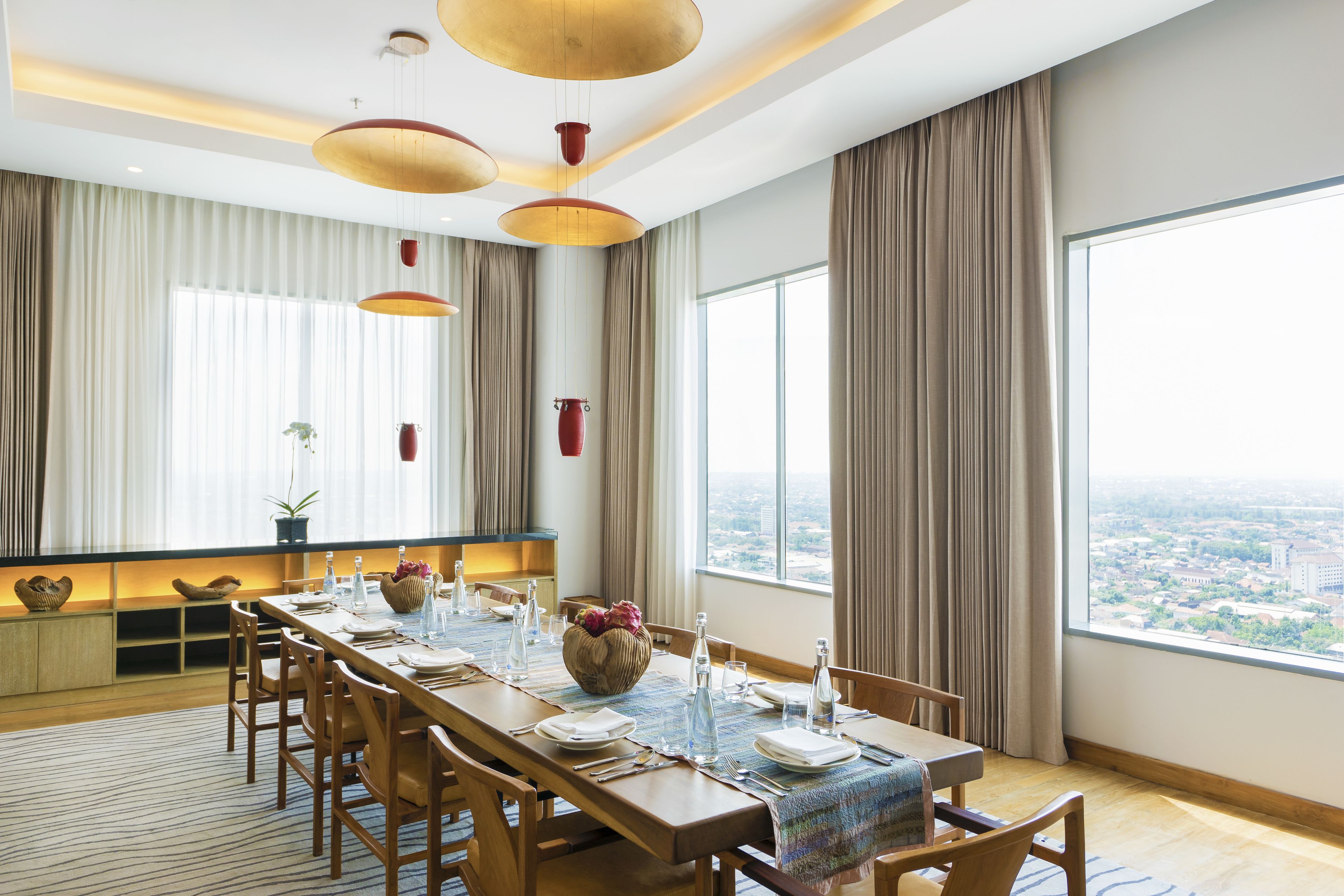 Un espace repas avec une longue table dressée pour six personnes, de grandes fenêtres avec des rideaux et des lampes suspendues.