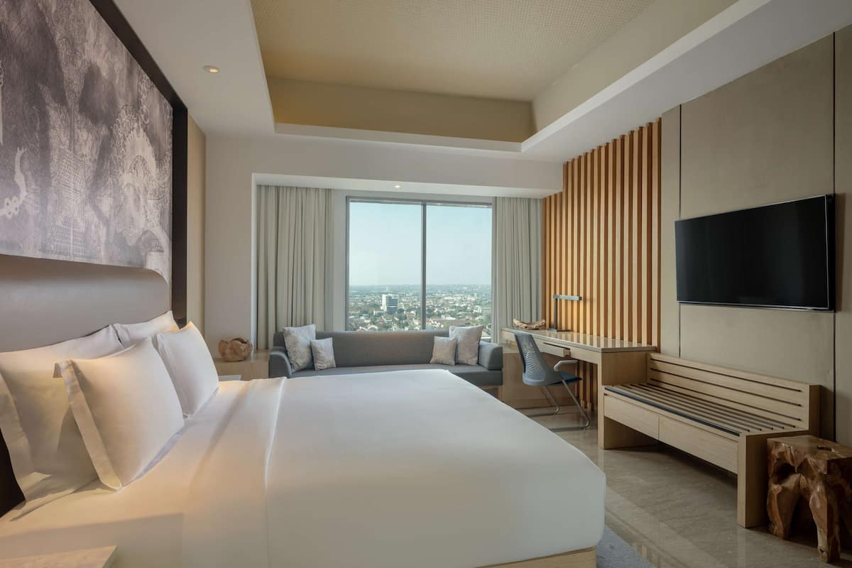 A modern hotel room with a large bed, a desk, a chair, a TV, and a view of the city.
