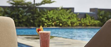 Un vaso de smoothie con adorno de fruta sobre una mesa junto a una silla de descanso al lado de la piscina.