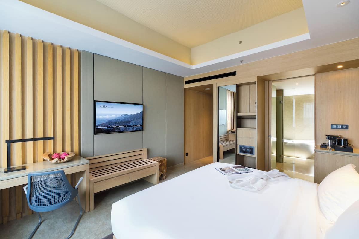 A modern hotel room with a large bed, a desk with a chair, a television, and a bathroom visible through an open door.