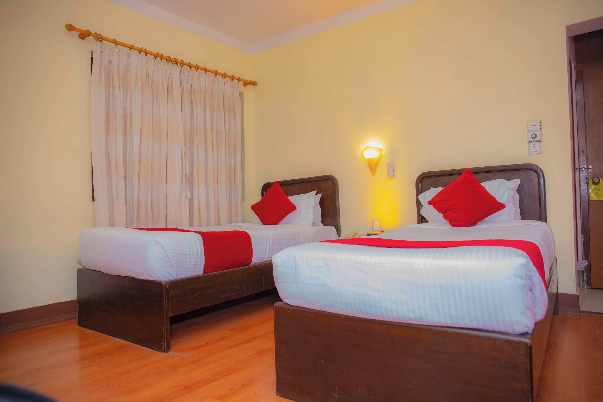 Een hotelkamer met twee bedden, houten hoofdeinden, een nachtkastje met een lamp, een raam met gordijnen en een deur.