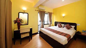 Quarto de hotel com uma cama grande, uma mesa de cabeceira com um vaso de flores, um espelho e uma janela com cortinas.
