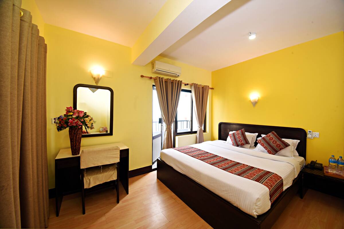 A hotel room with a large bed, a bedside table with a vase of flowers, a mirror, and a window with curtains.
