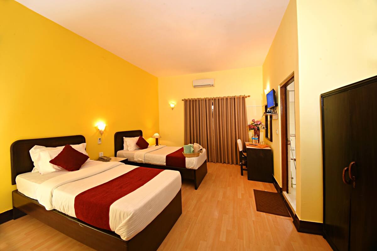 A hotel room with two beds, a desk, a wardrobe, and an air conditioning unit.