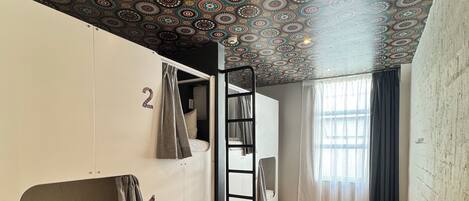 A dormitory room with bunk beds, a ladder, a window with curtains, and a patterned ceiling.