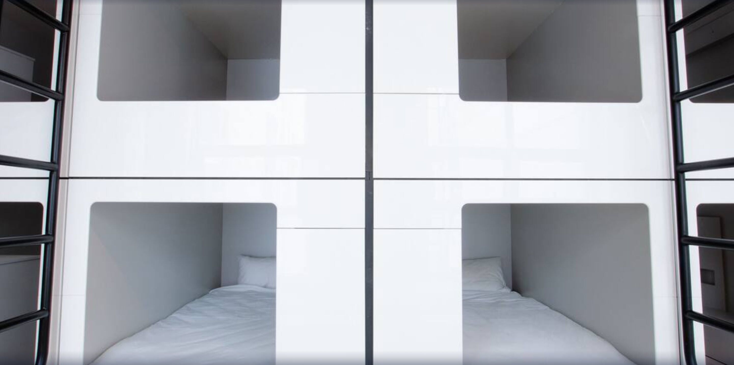 Quarto de hotel moderno com design minimalista, apresentando quatro camas dispostas em padrão quadrado, cada uma com colchão branco e almofadas combinando.