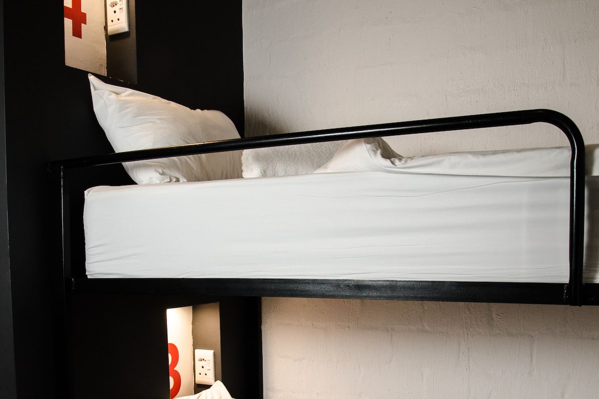 Una cama litera con ropa de cama blanca y estructura de metal negro. En la pared hay números iluminados que indican los números de habitación.