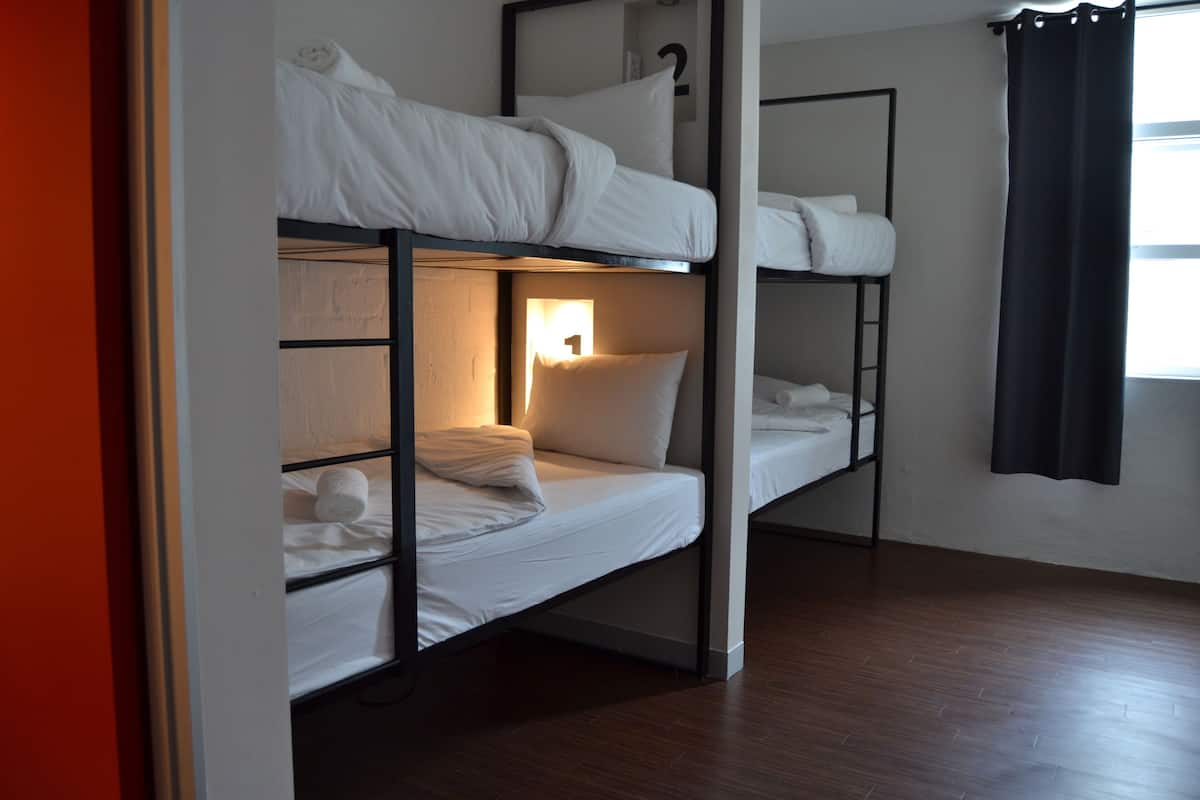 Habitación con literas, un cartel con un número en la pared, suelo de madera y una ventana con cortina.