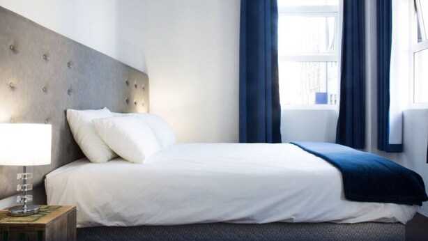 A neatly made bed with white linens and a blue throw blanket, a bedside lamp, and a view of the outside through a window with blue curtains. - 91 Loop - Hostel (Cape Town)