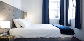 Een net opgemaakt bed met witte lakens en een blauwe plaid, een bedlampje en uitzicht naar buiten door een raam met blauwe gordijnen.