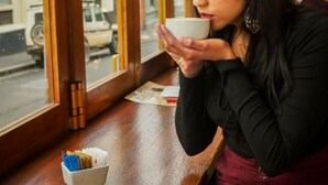 A person sitting at a wooden table inside a cafe with a window view of the street outside. - 91 Loop - Hostel (Cape Town)