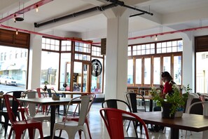 Una cafetería con amplios ventanales, sillas rojas y blancas, y una persona trabajando en una mesa.