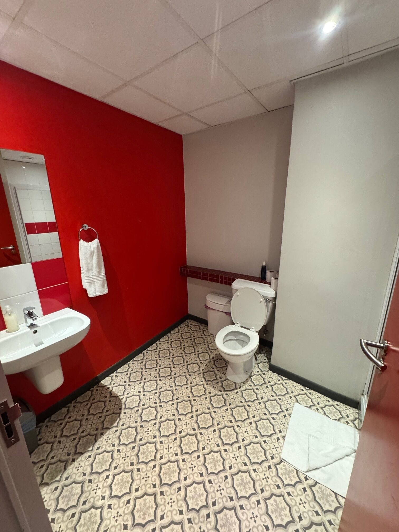 Une salle de bain avec un mur rouge, un lavabo blanc, des toilettes et un sol à motifs.