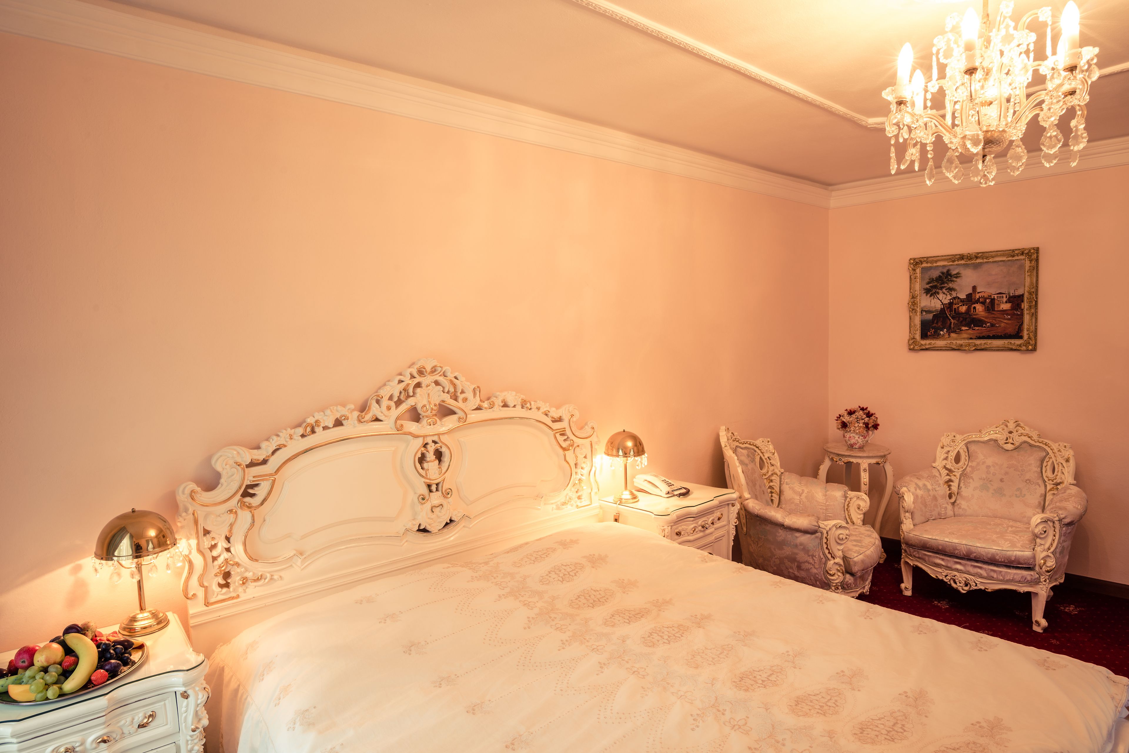Un dormitorio con una cama grande y ornamentada, dos sillones antiguos, una mesita de noche con lámpara y un cuadro en la pared.