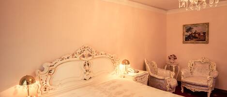Un dormitorio con una cama grande y ornamentada, dos sillones antiguos, una mesita de noche con lámpara y un cuadro en la pared.