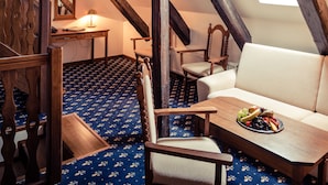 Een zolderkamer met een schuine plafond, houten balken, een bank, een tafel met fruit en een kamerbreed tapijt met dessin.