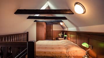 Um quarto com teto inclinado, uma cama com cabeceira de madeira, uma mesinha de cabeceira com abajur e uma pequena mesa com uma planta.