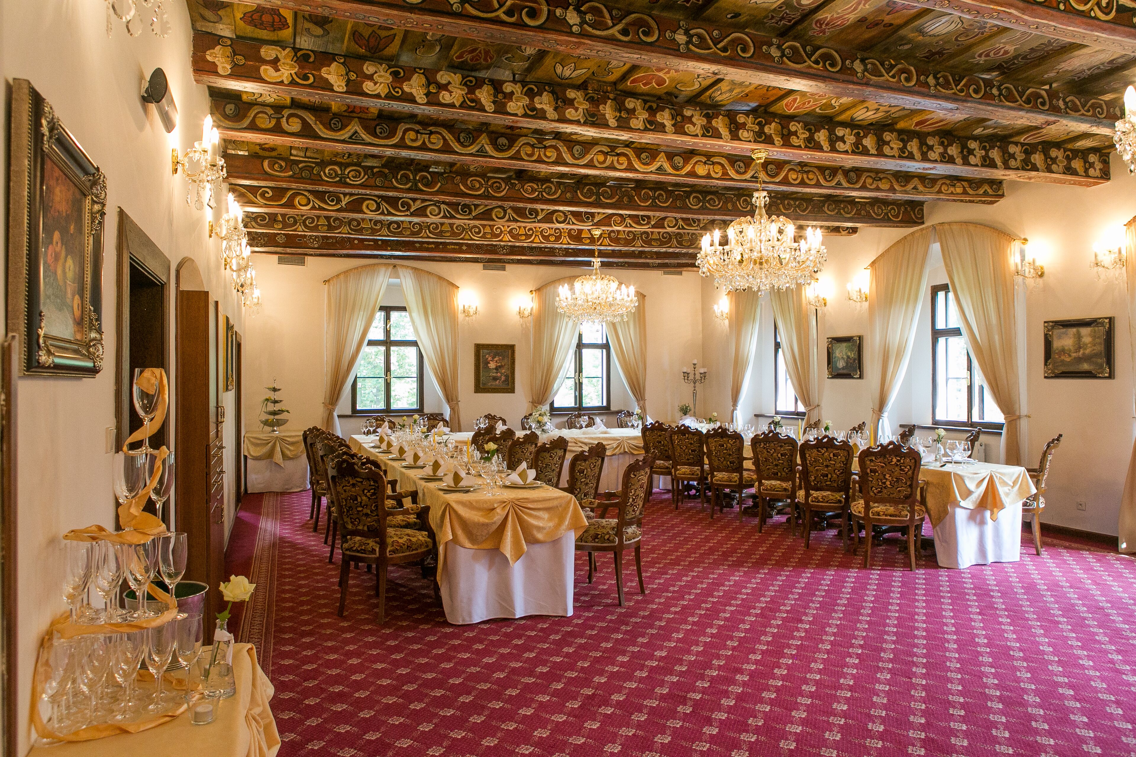 Een formele eetzaal met een versierde plafond, kroonluchters en een rode vloerbedekking.