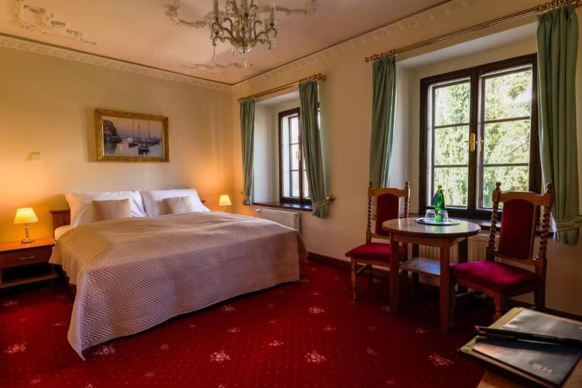 Habitación de hotel con cama, dos sillas, una mesa, candelabro, ventana con cortinas y un cuadro en la pared.