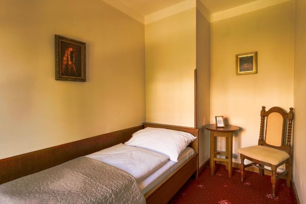 Een enkel bed met een houten hoofdbord, een klein houten stoeltje en twee ingelijste foto's aan de muur.