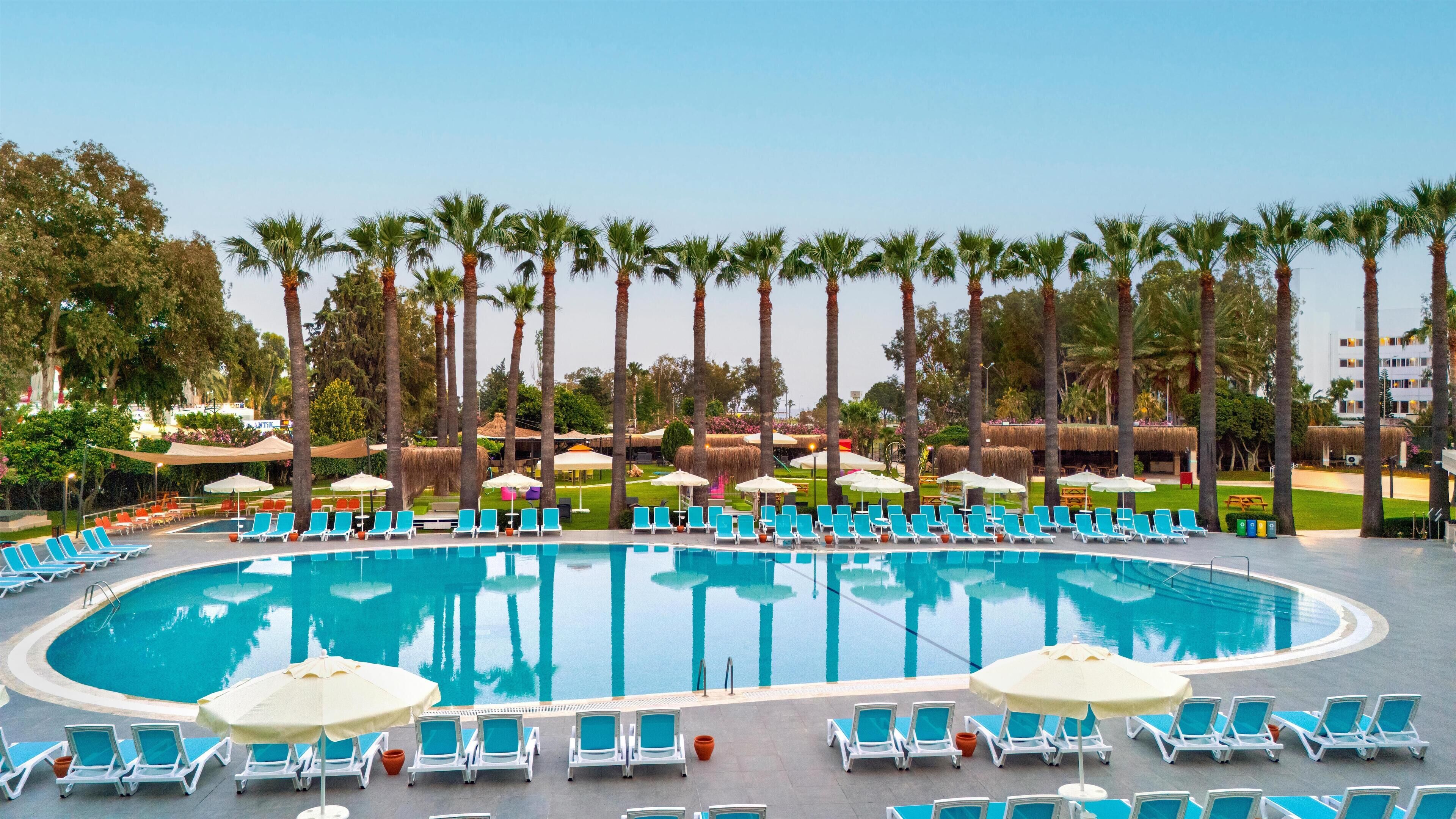 A large oval pool surrounded by lounge chairs and umbrellas, with palm trees and a thatched-roof structure in the background.