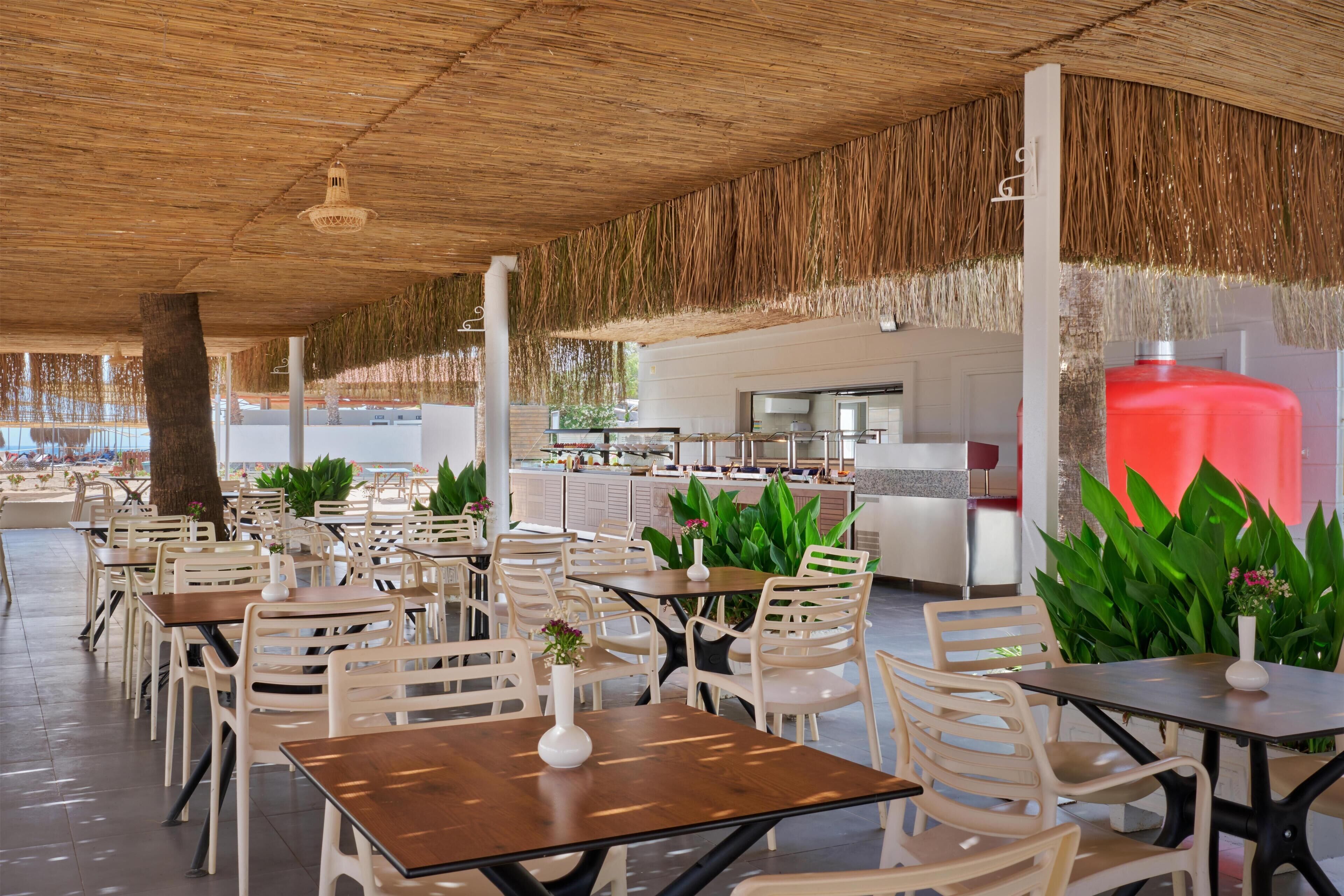 A spacious outdoor dining area with multiple tables and chairs, a central bar area, and lush green plants.