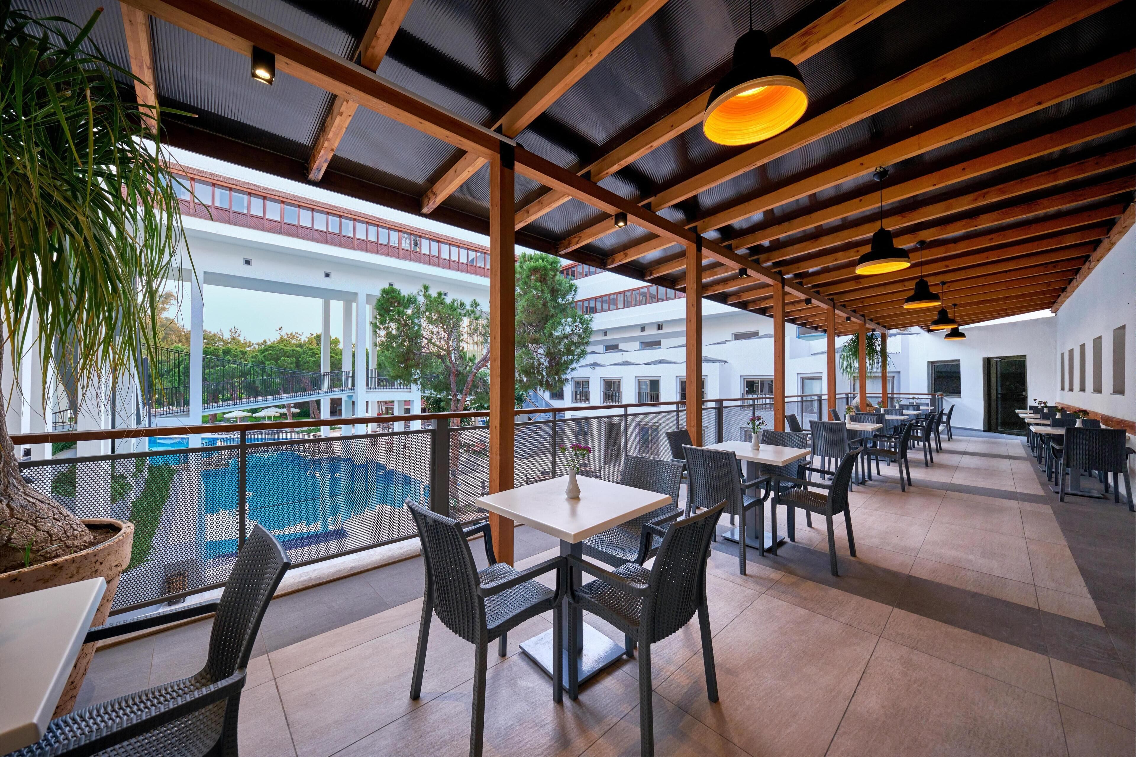 Une terrasse extérieure avec des tables et des chaises, donnant sur une piscine et des bâtiments.