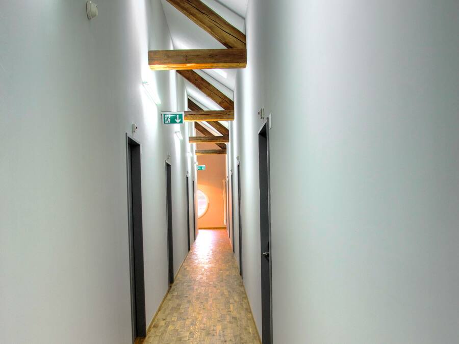 A long, narrow hallway with wooden flooring and white walls, featuring ceiling beams and doors on both sides.