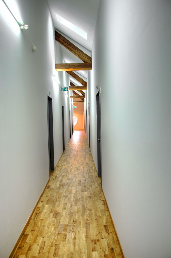 A long, narrow hallway with wooden flooring and white walls, featuring ceiling beams and doors on both sides.