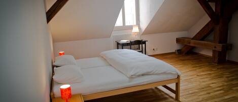 Un dormitorio con cama de madera, una mesita de noche con una lámpara roja, una silla y una ventana con lámpara.