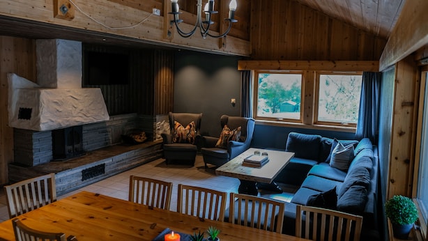 Un interior de cabaña acogedor, con una chimenea de piedra, una mesa de comedor de madera y un sofá junto a una ventana.