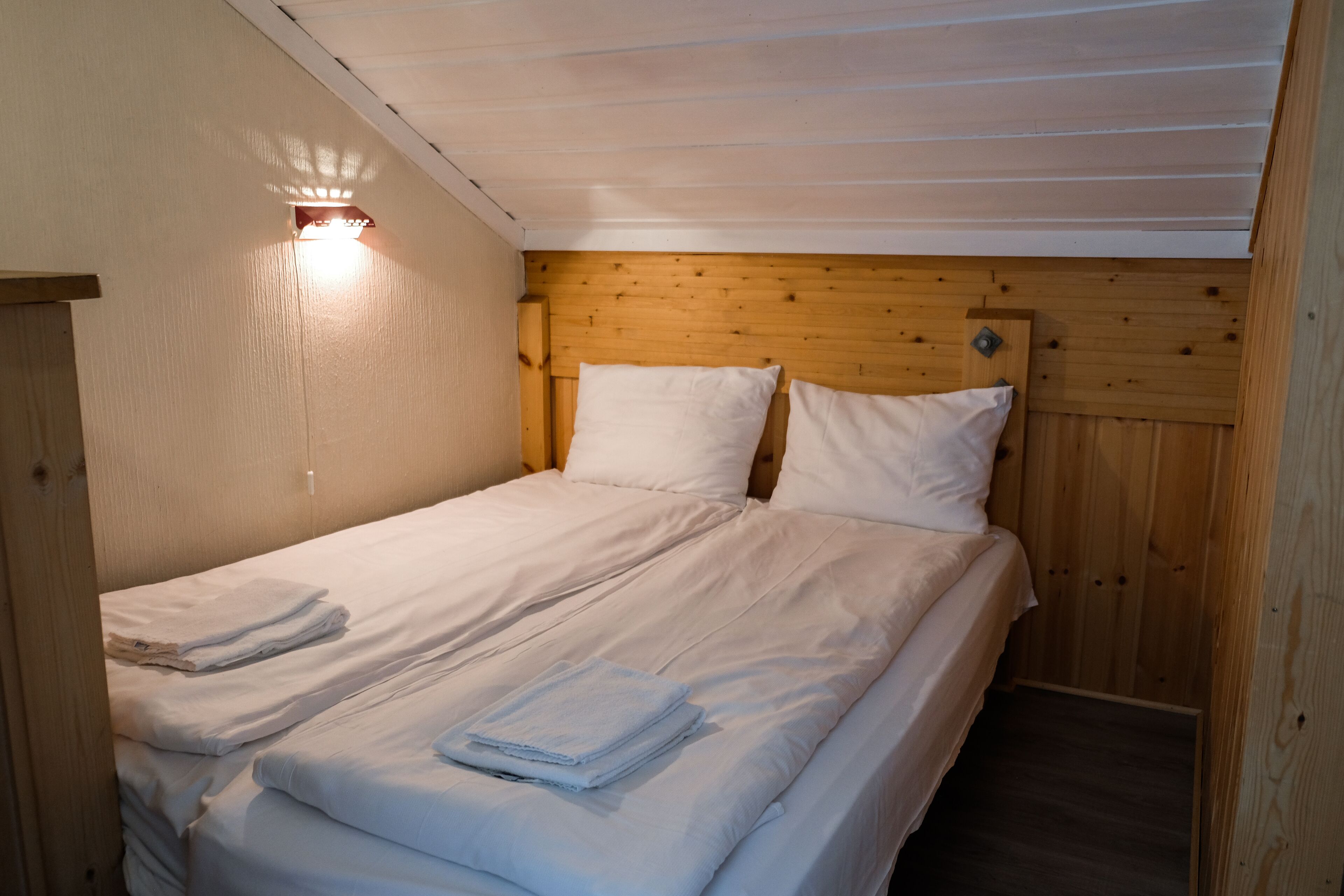 A single bed with white linens and pillows, a wooden headboard, and a wall-mounted light fixture.