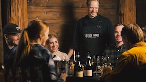 Un grupo de personas sentadas alrededor de una mesa con botellas y copas de vino, en una habitación con paredes de madera y una lámpara colgante.