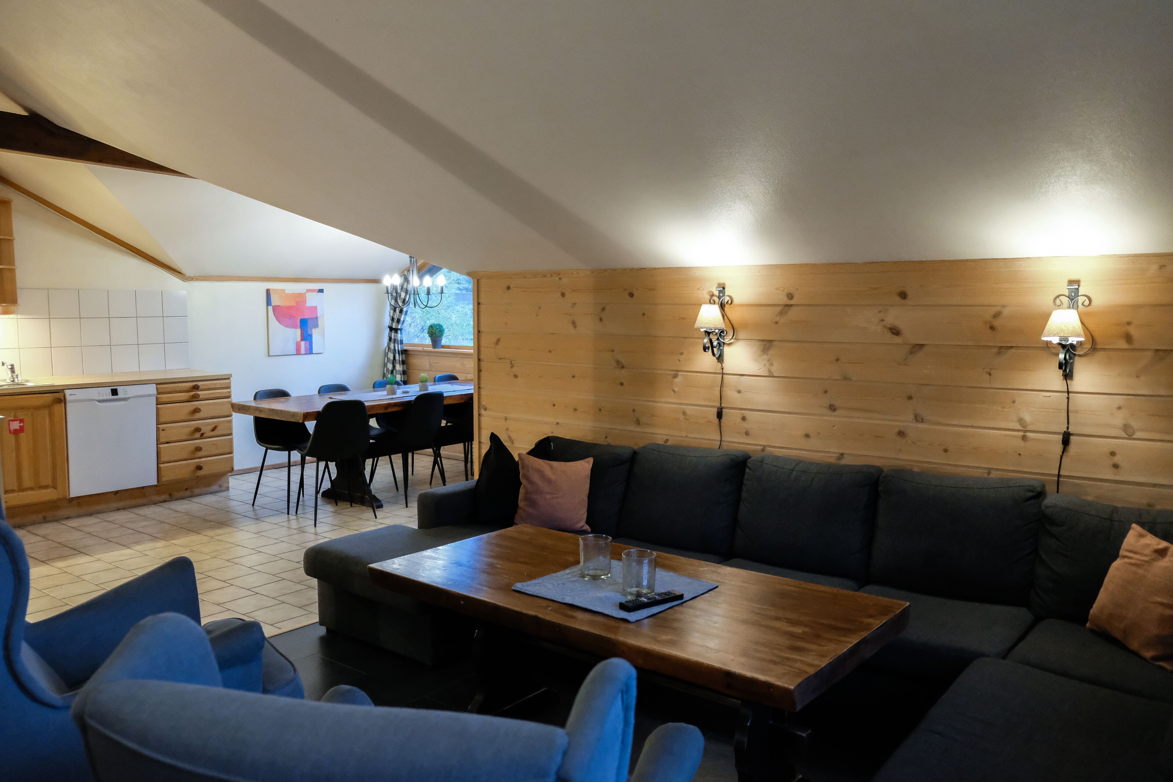 A cozy living area with a wooden dining table, blue chairs, a sofa, and a kitchen in the background.