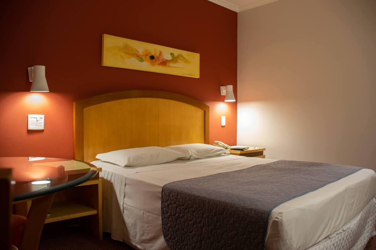 A hotel room with a wooden headboard, a round glass table, a bed with white bedding and a gray blanket, and two bedside lamps.