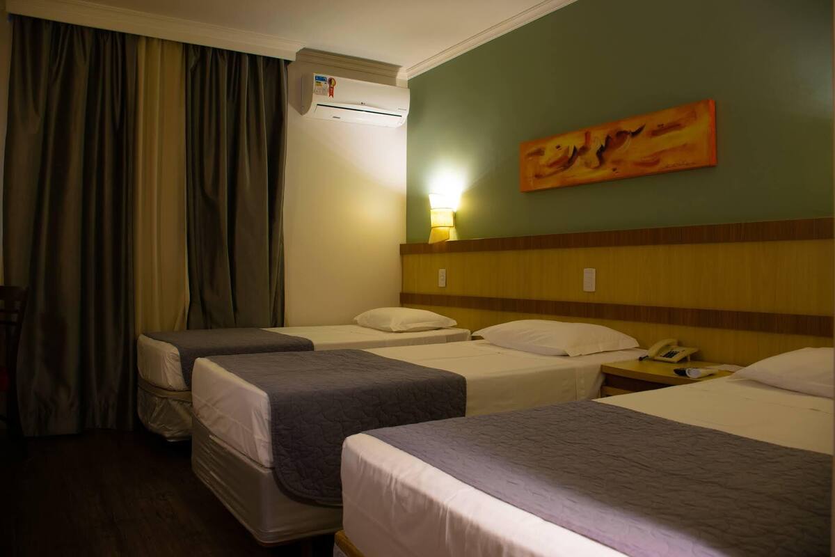 A hotel room with three beds, an air conditioning unit, a painting on the wall, and a nightstand with a phone.