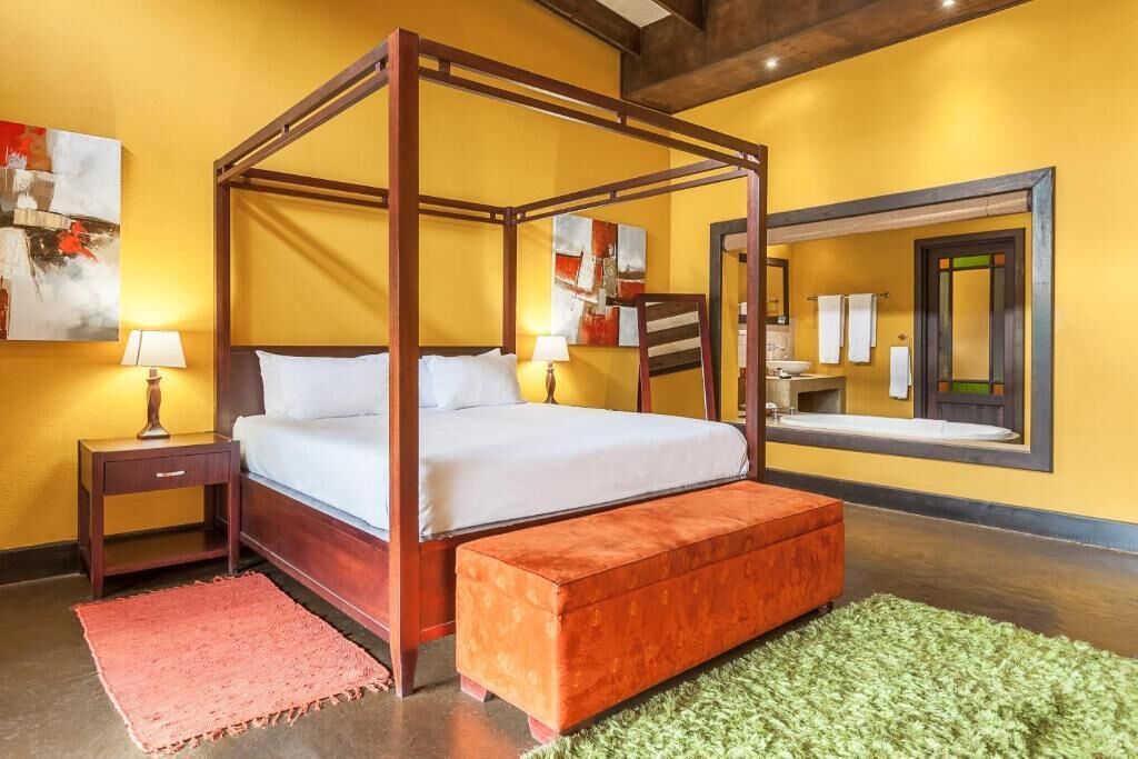 A four-poster bed with a wooden frame, a green rug, and a red ottoman in a room with yellow walls and a large mirror.
