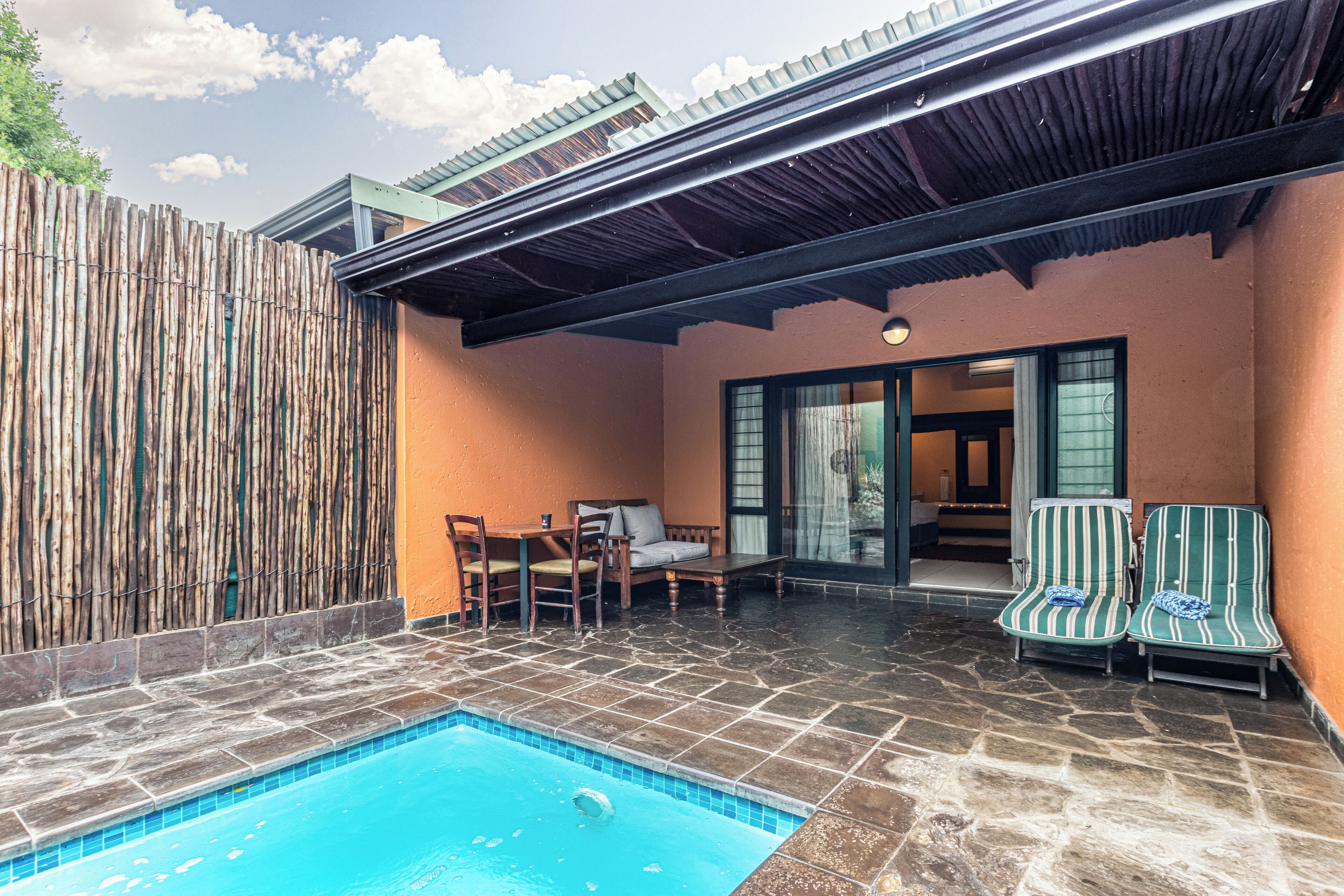 A pool area with stone flooring, a wooden fence, and a covered seating area with chairs and a table.