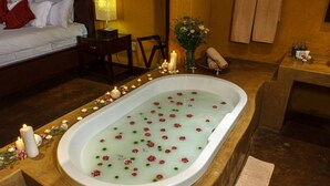 Una habitación con una bañera grande llena de agua y flores, una cama con ropa blanca, una mesita de noche de madera, y una pequeña mesa con velas y un florero.