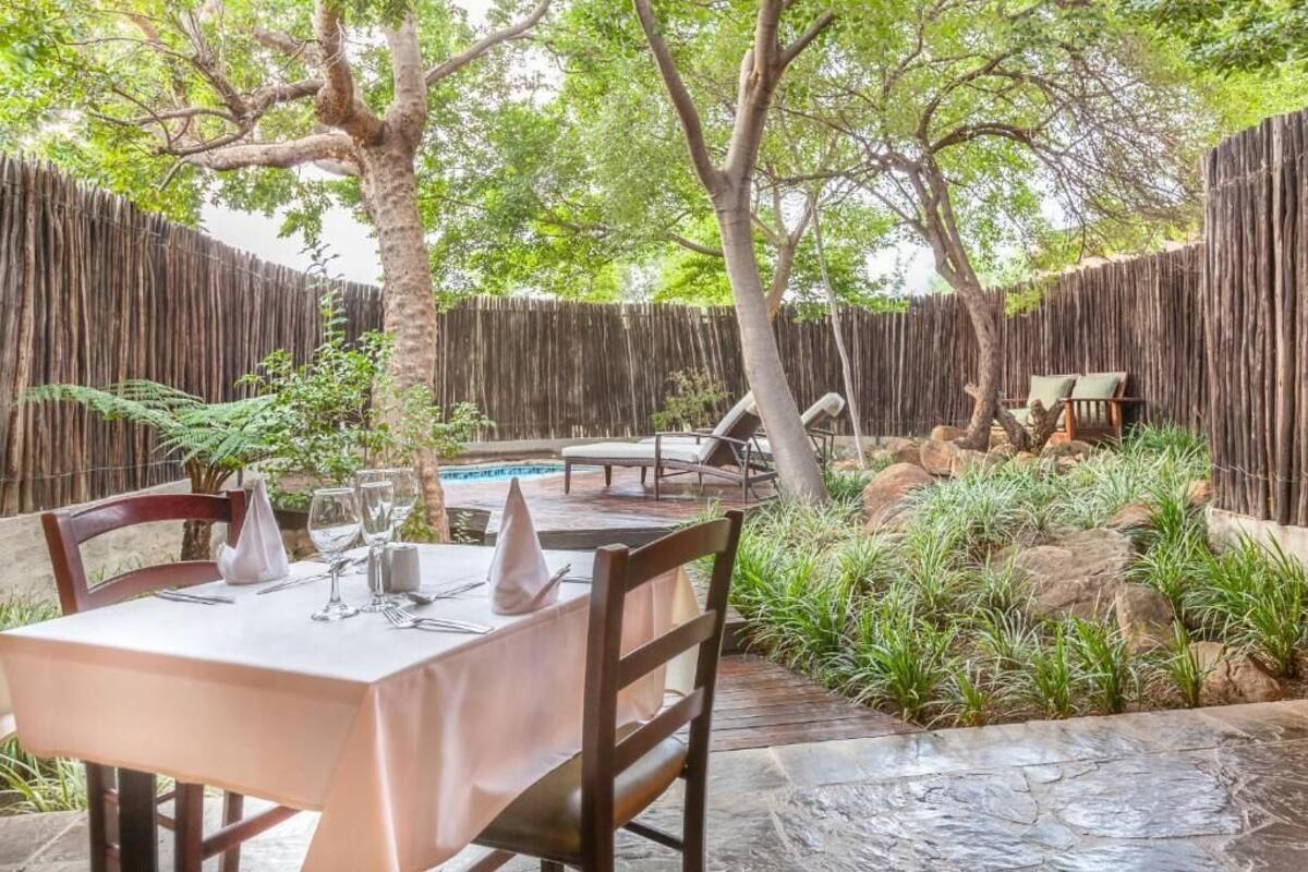 An outdoor dining area with a table set for two, surrounded by a wooden fence and lush greenery.