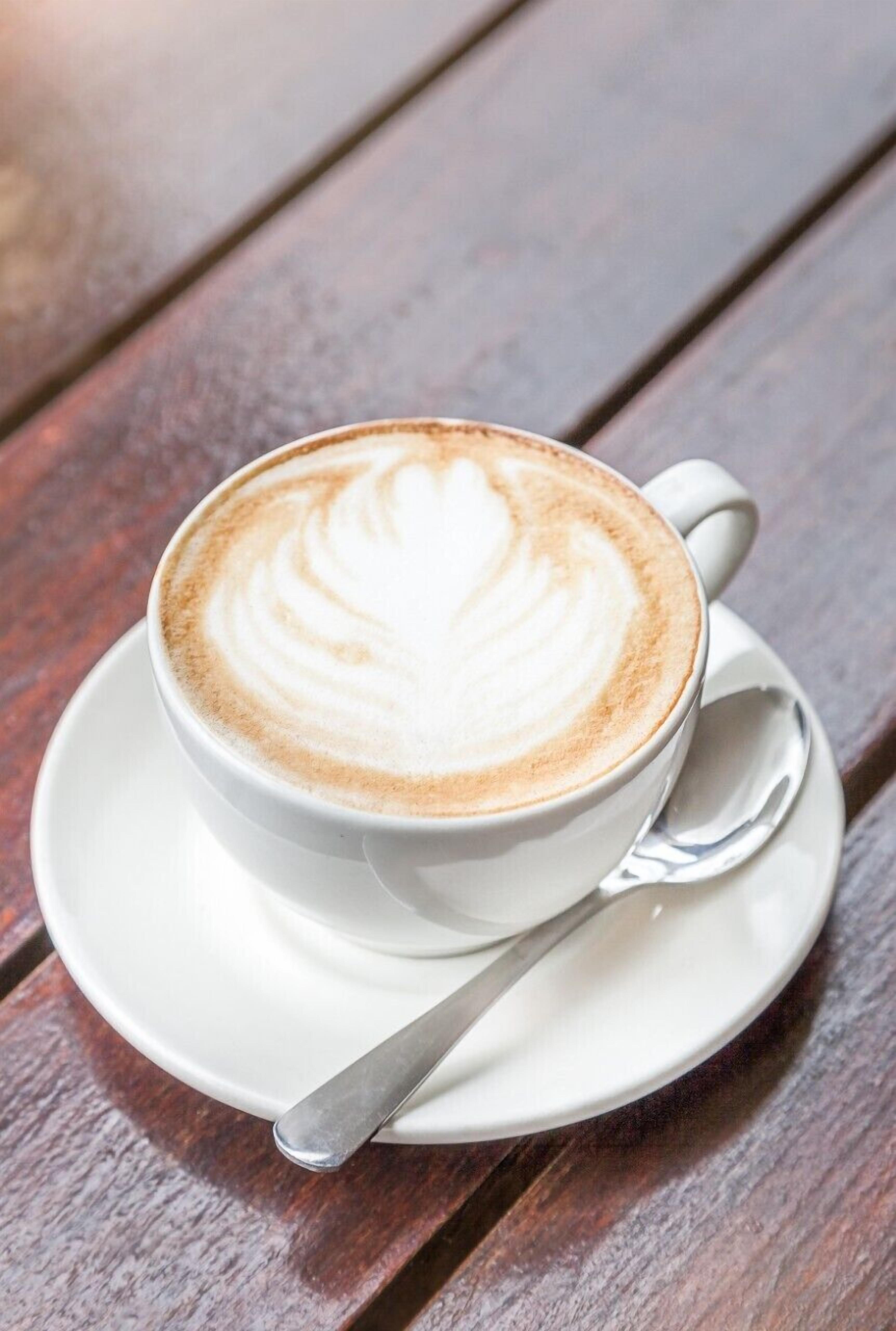 A cup of coffee with latte art on a white saucer and spoon, placed on a wooden table.