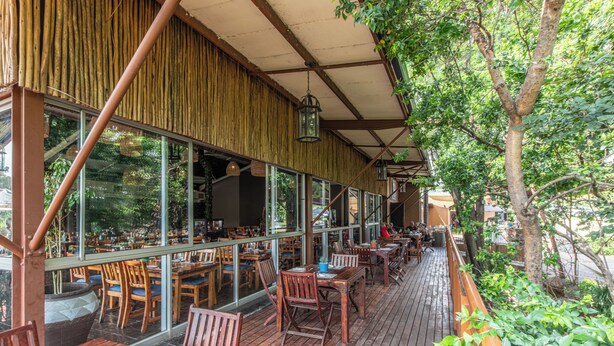 Un restaurante al aire libre con mesas y sillas de madera, ventanas amplias y una zona cubierta con pared de bambú.