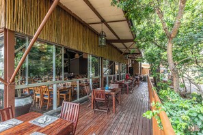 Un restaurante al aire libre con mesas y sillas de madera, ventanas amplias y una zona cubierta con pared de bambú.
