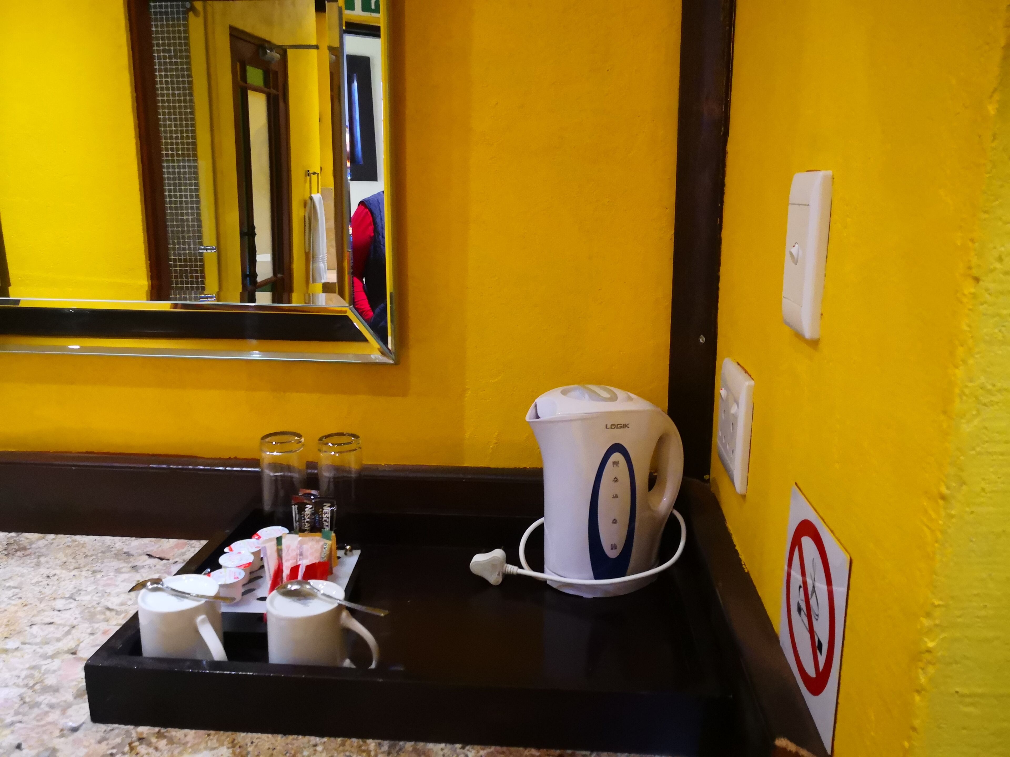 A hotel room with a yellow wall, a black countertop, a white kettle, and a tray with glasses and a mug.
